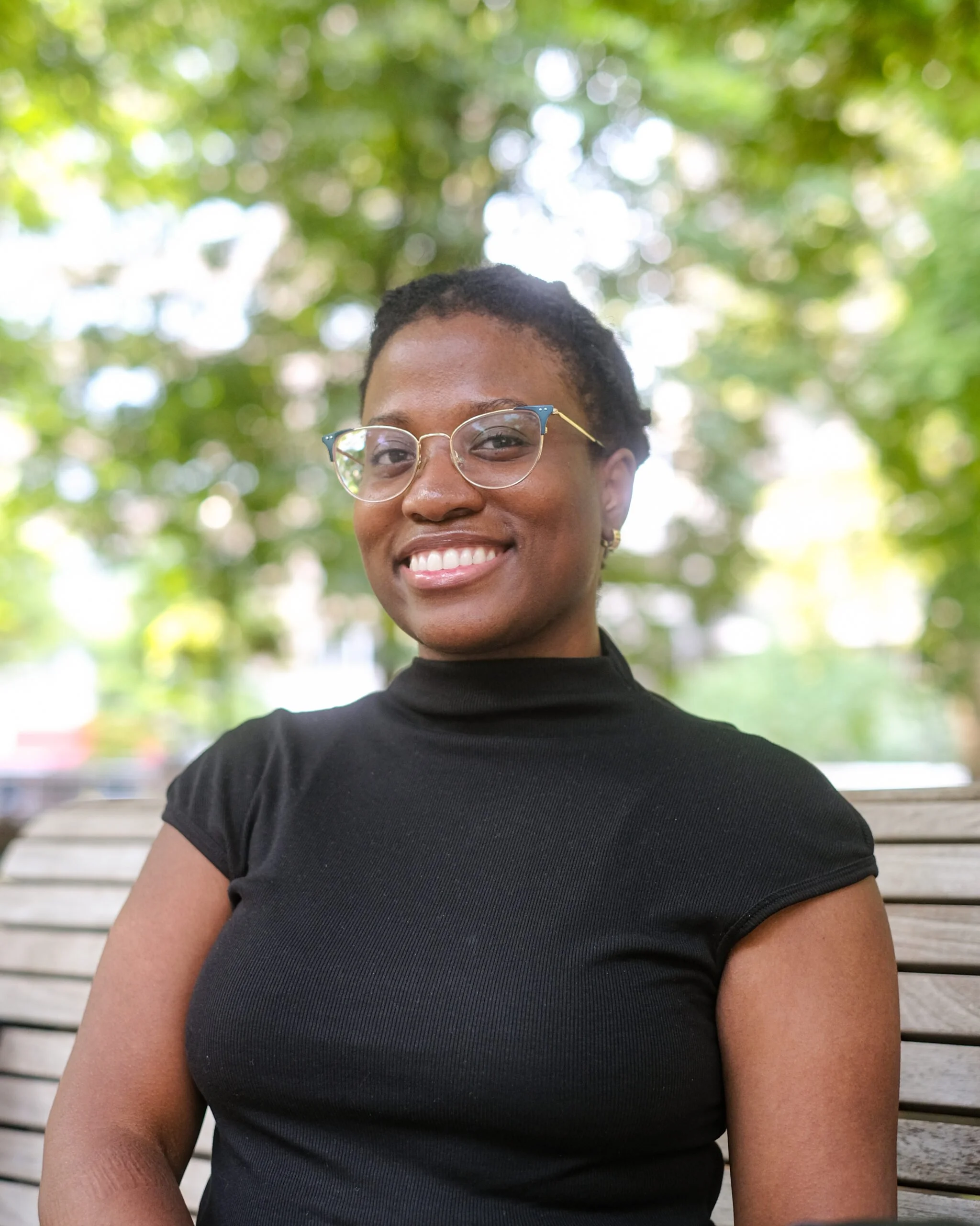 Portrait of a smiling woman with short curly hair and glasses, wearing a black top, sitting on a park bench with green trees in the background.