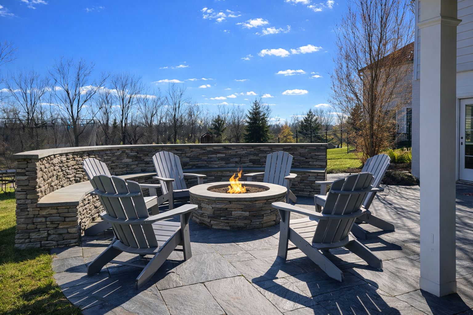 Outdoor patio with a circular fire pit and six Adirondack chairs on a stone-paved surface, surrounded by a stone retaining wall, with trees and a house in the background under a blue sky.