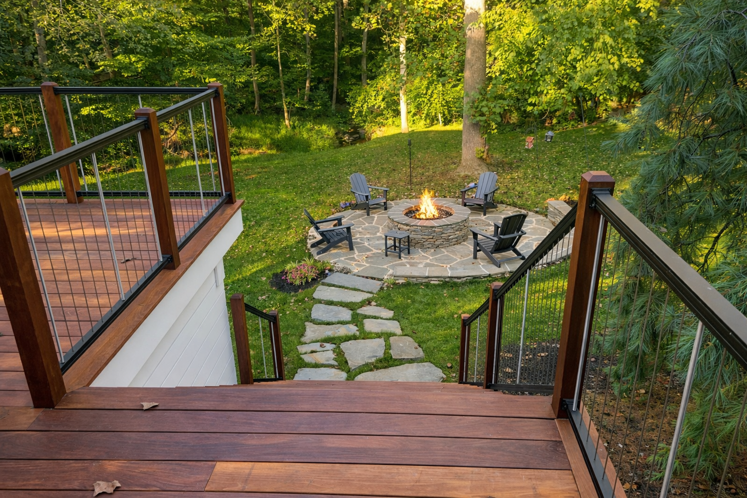 View from a wooden deck overlooking a backyard with a stone fire pit surrounded by Adirondack chairs, tall green trees, and lush grass.