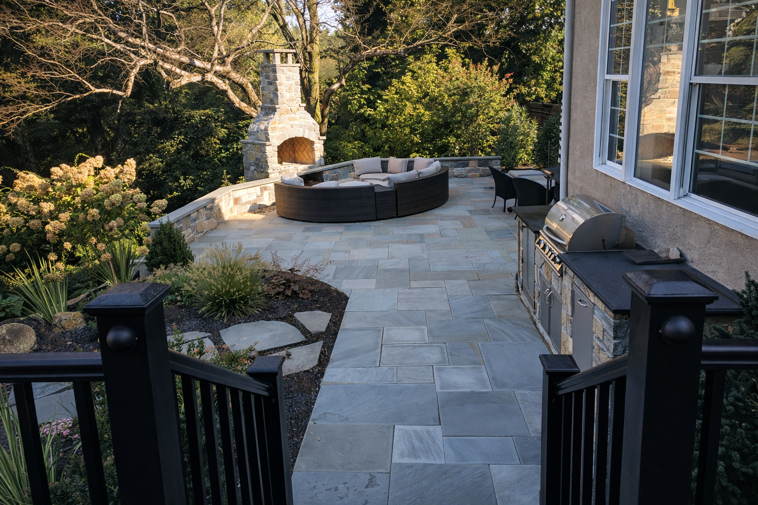 A spacious outdoor patio with stone flooring, a built-in stone fireplace, garden plants on the left, a sectional outdoor sofa, a small dining table with chairs, and a built-in grill on the right, surrounded by trees and greenery.