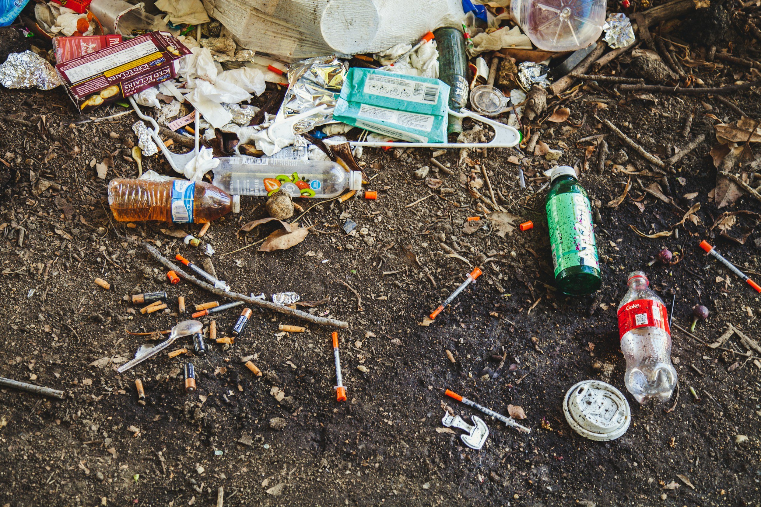 Litter and trash scattered on the dirt ground, including cigarette butts, plastic bottles, aluminum cans, food wrappers, a disposable mask, and small debris.