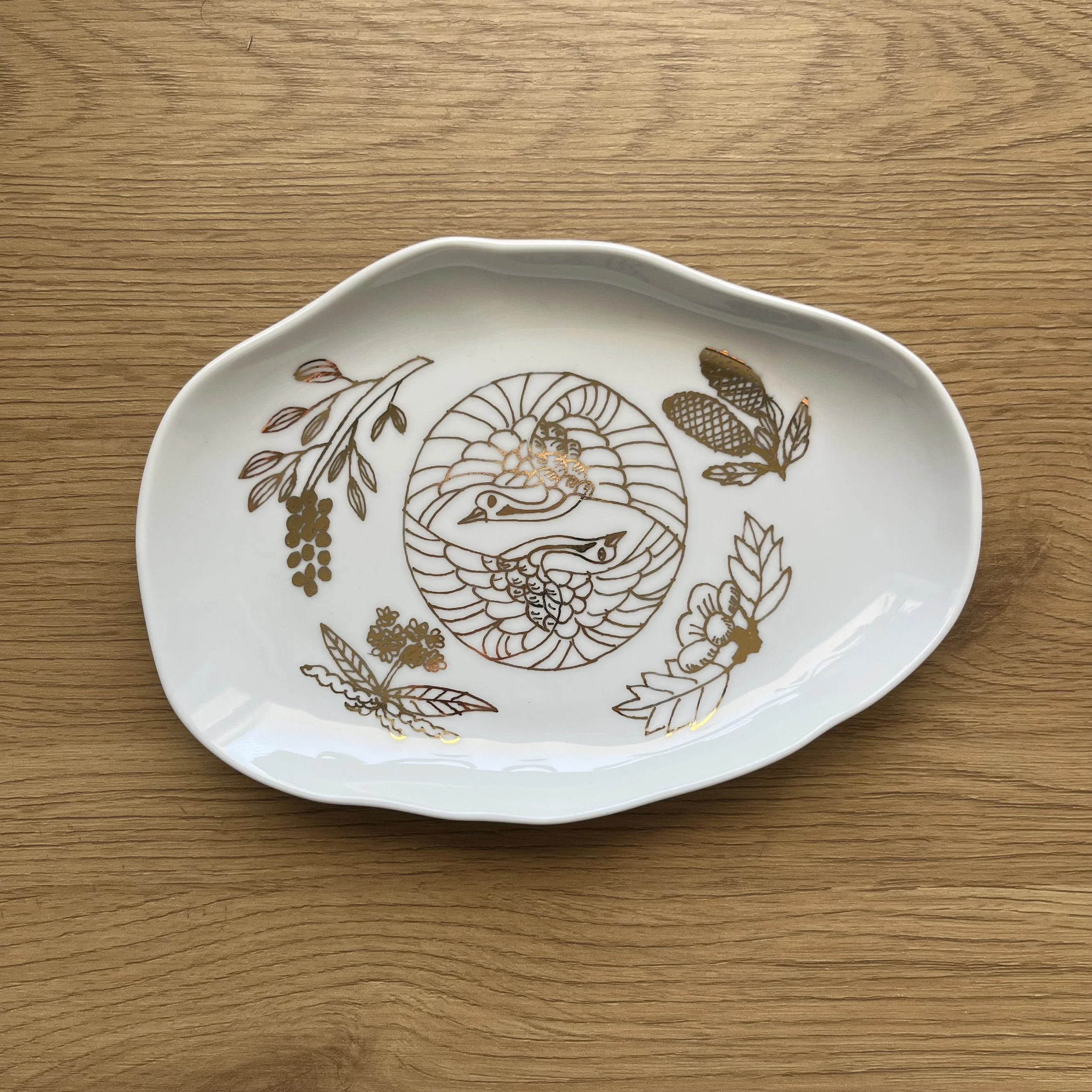 A white ceramic dish with gold decorative pattern featuring two swans and various botanical motifs, placed on a wooden surface.