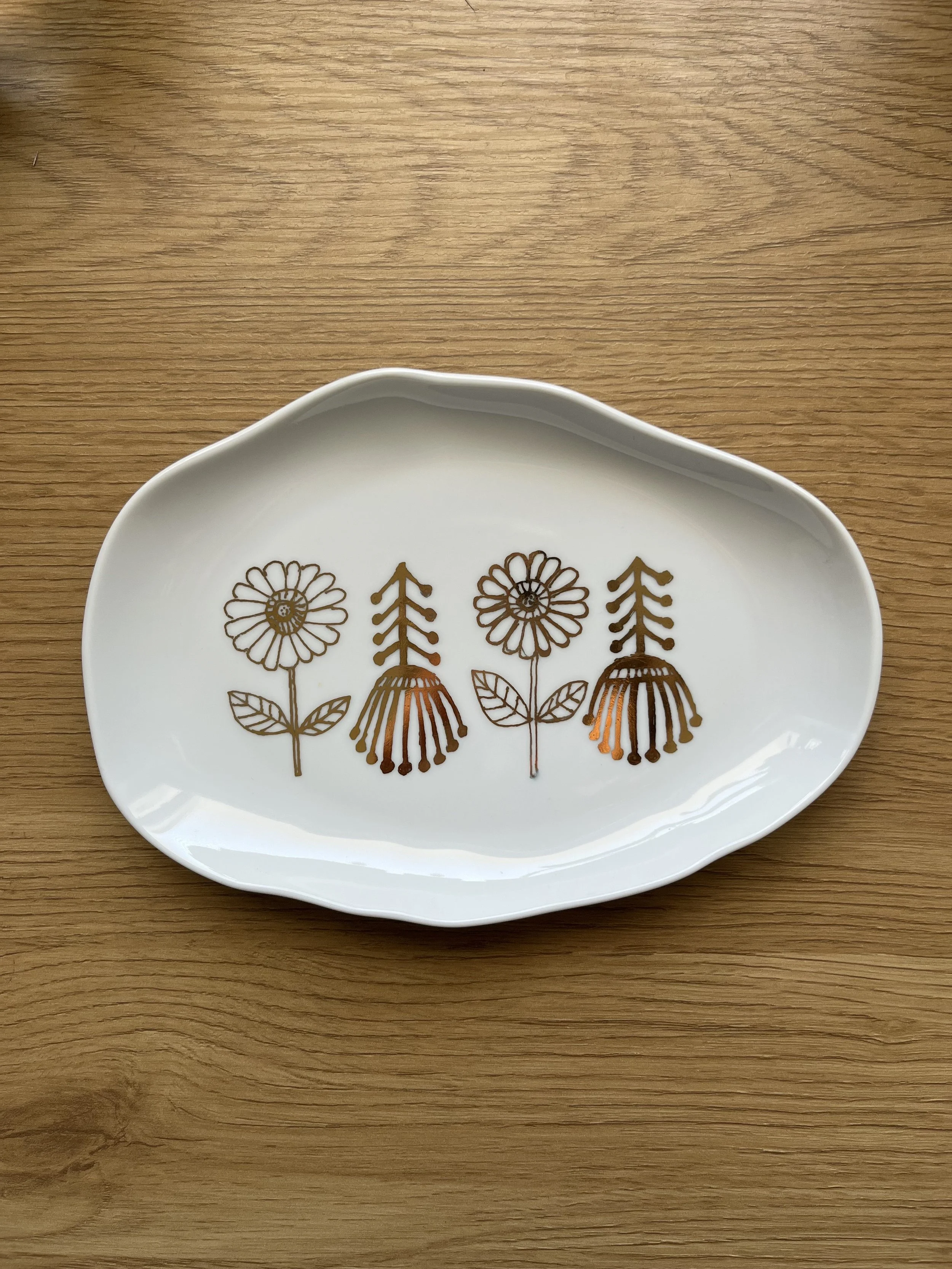 White oval dish with a floral pattern in gold featuring three flowers with leaves, placed on a wooden surface.