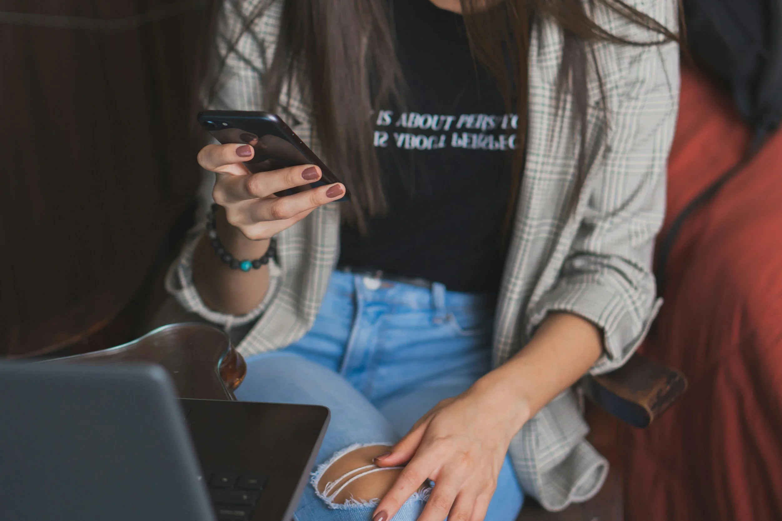 A woman sitting on a couch using her smartphone, wearing a plaid blazer, ripped jeans, and a black t-shirt.