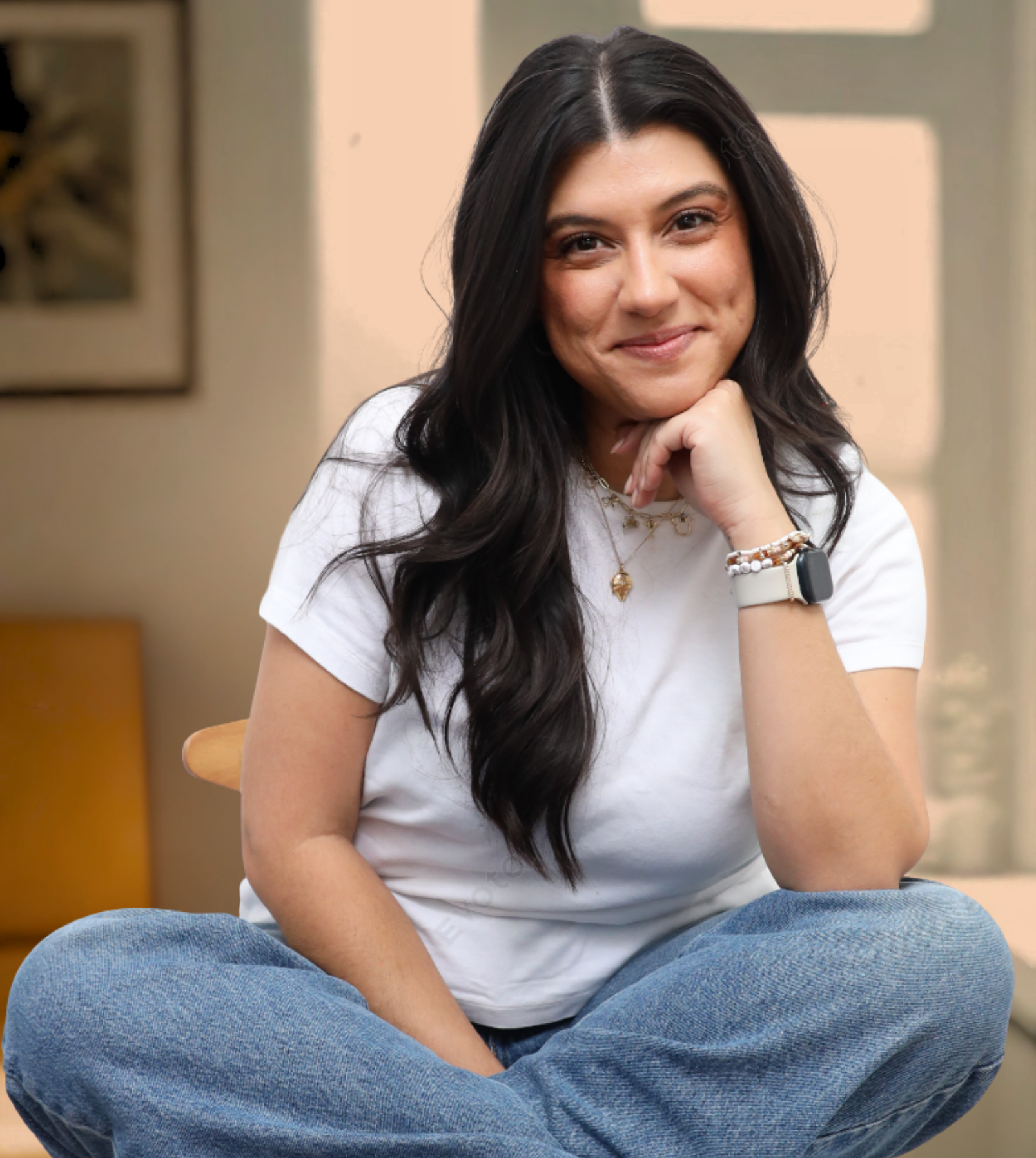 A woman with long black hair, wearing a white t-shirt and blue jeans, sitting cross-legged on a chair with her chin resting on her hand, smiling softly at the camera.