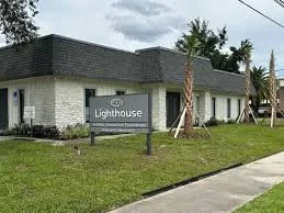 A building with a sign that reads 'Lighthouse,' surrounded by a grassy area, palm trees, and a sidewalk.