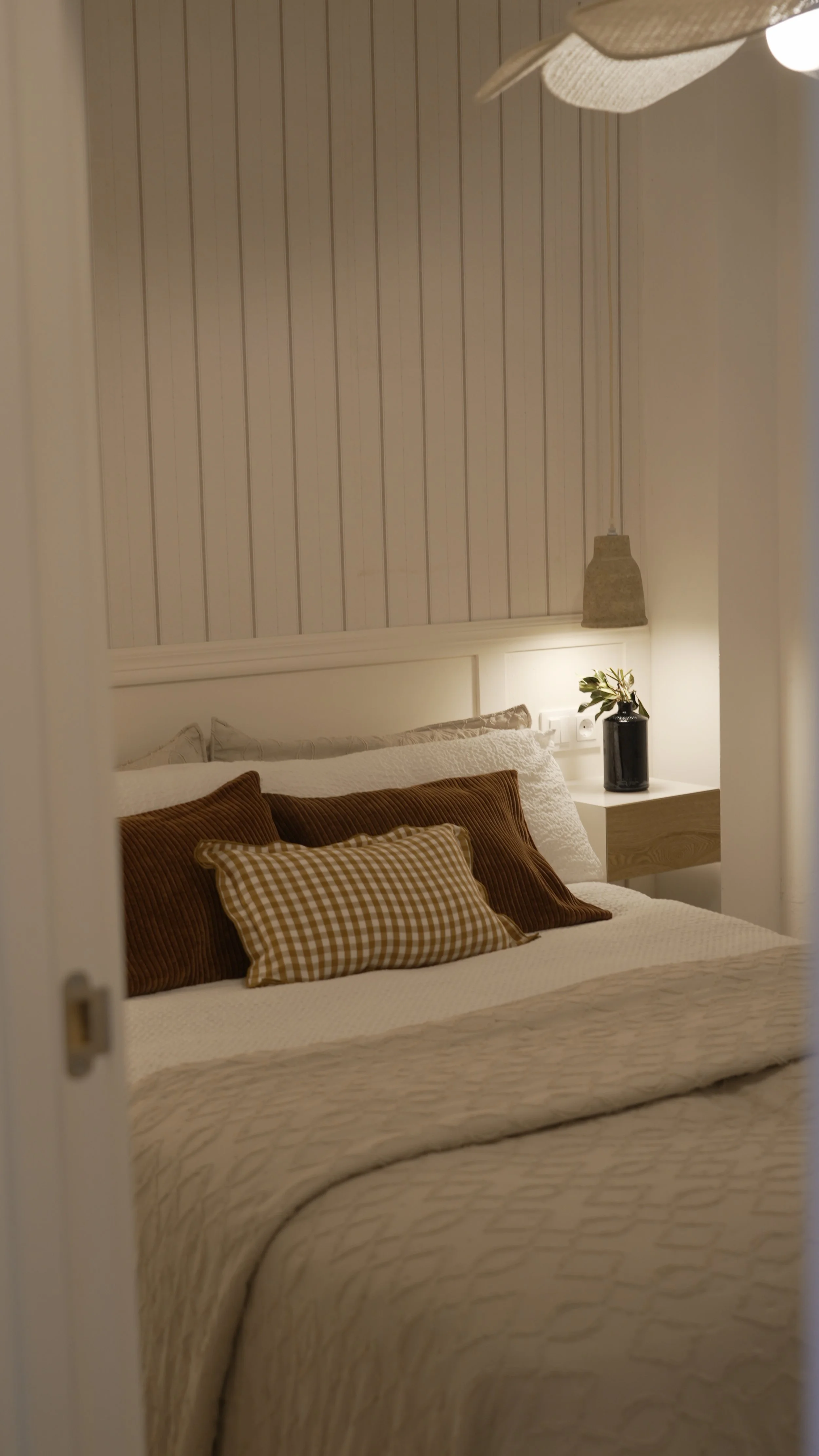 Cama en una habitación decorada en tonos neutros, con almohadas color marrón y cachemira, y un velador con planta junto a una lámpara de techo.