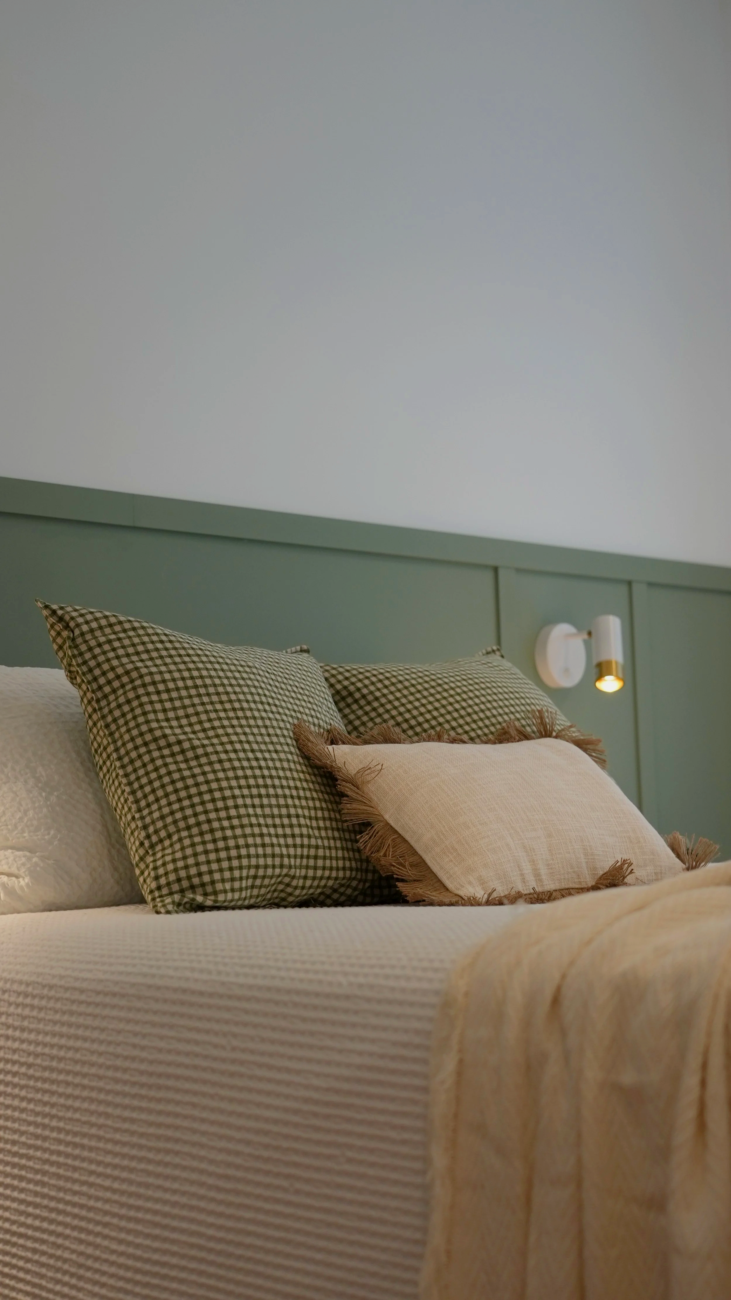 Cama con almohadas de tejidos diferentes, la cabecera es de color verde y hay una luz de lectura en la pared de fondo.