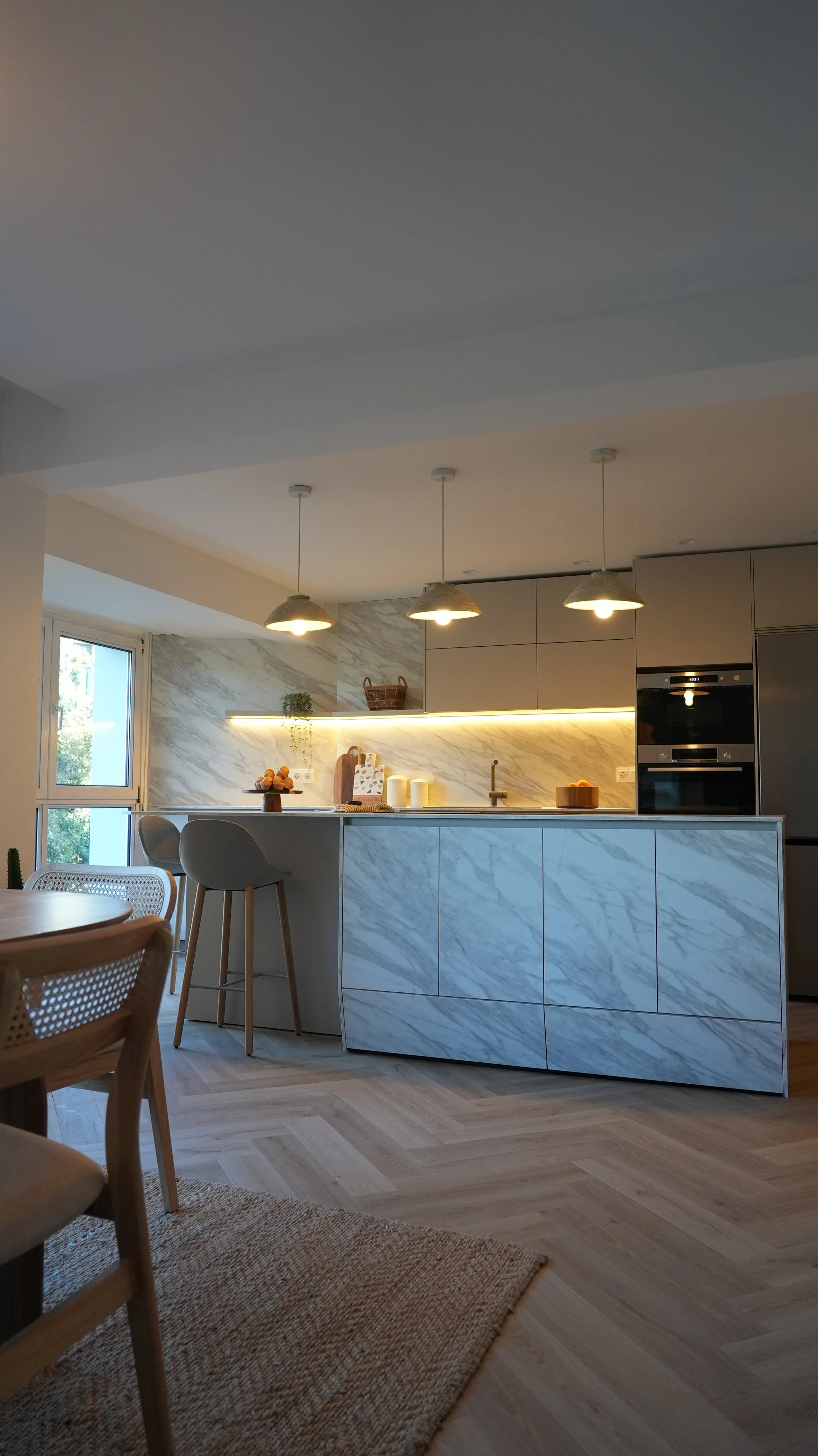 Cocina moderna con encimera y gavetas de mármol, iluminación empotrada y luminarias colgantes sobre la isla, cerca de una ventana que deja entrar luz natural.
