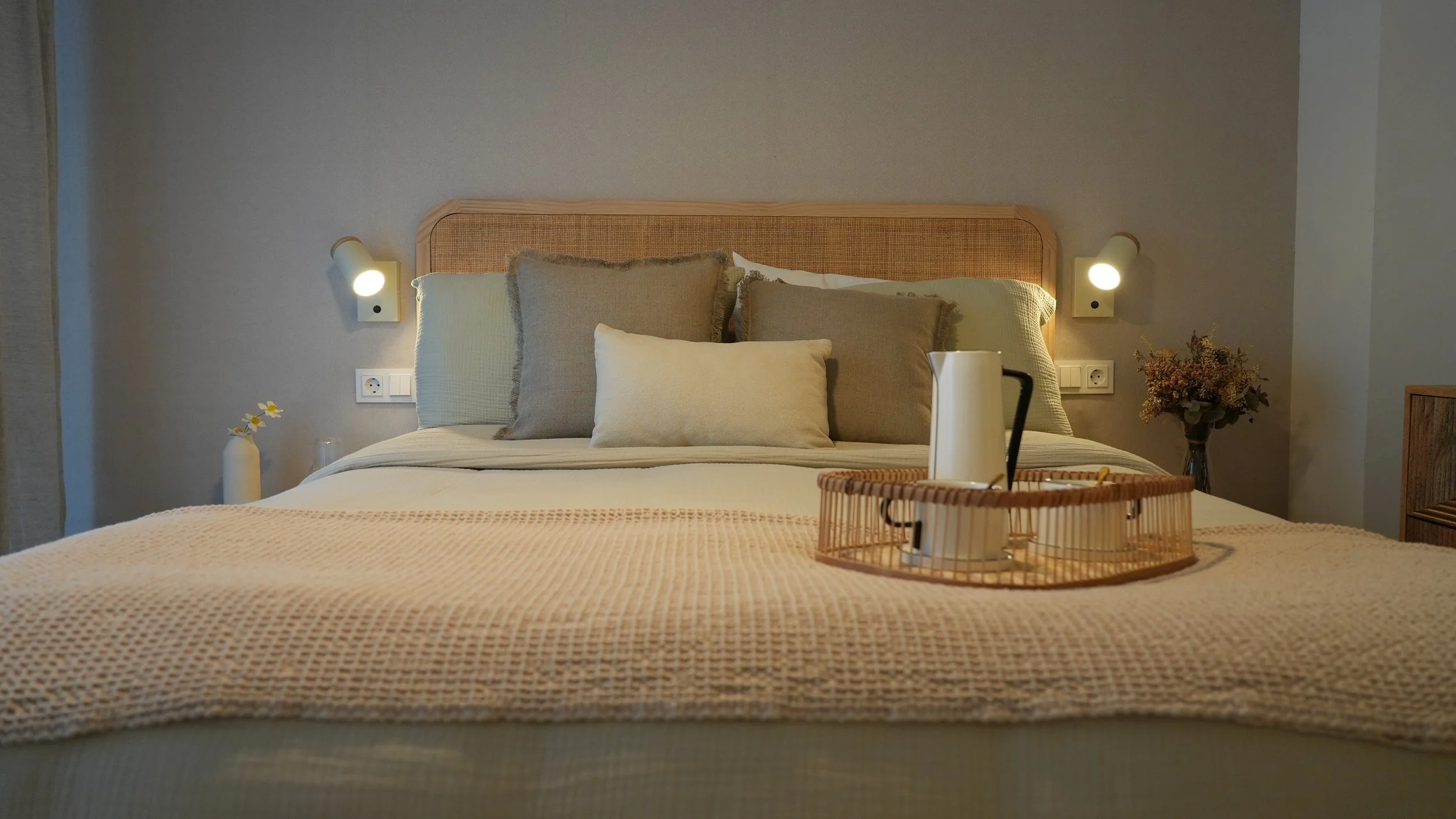 Cama de habitación con ropa de cama en tonos neutros, luz suave y decoración minimalista con flores y utensilios de desayuno en bandeja.