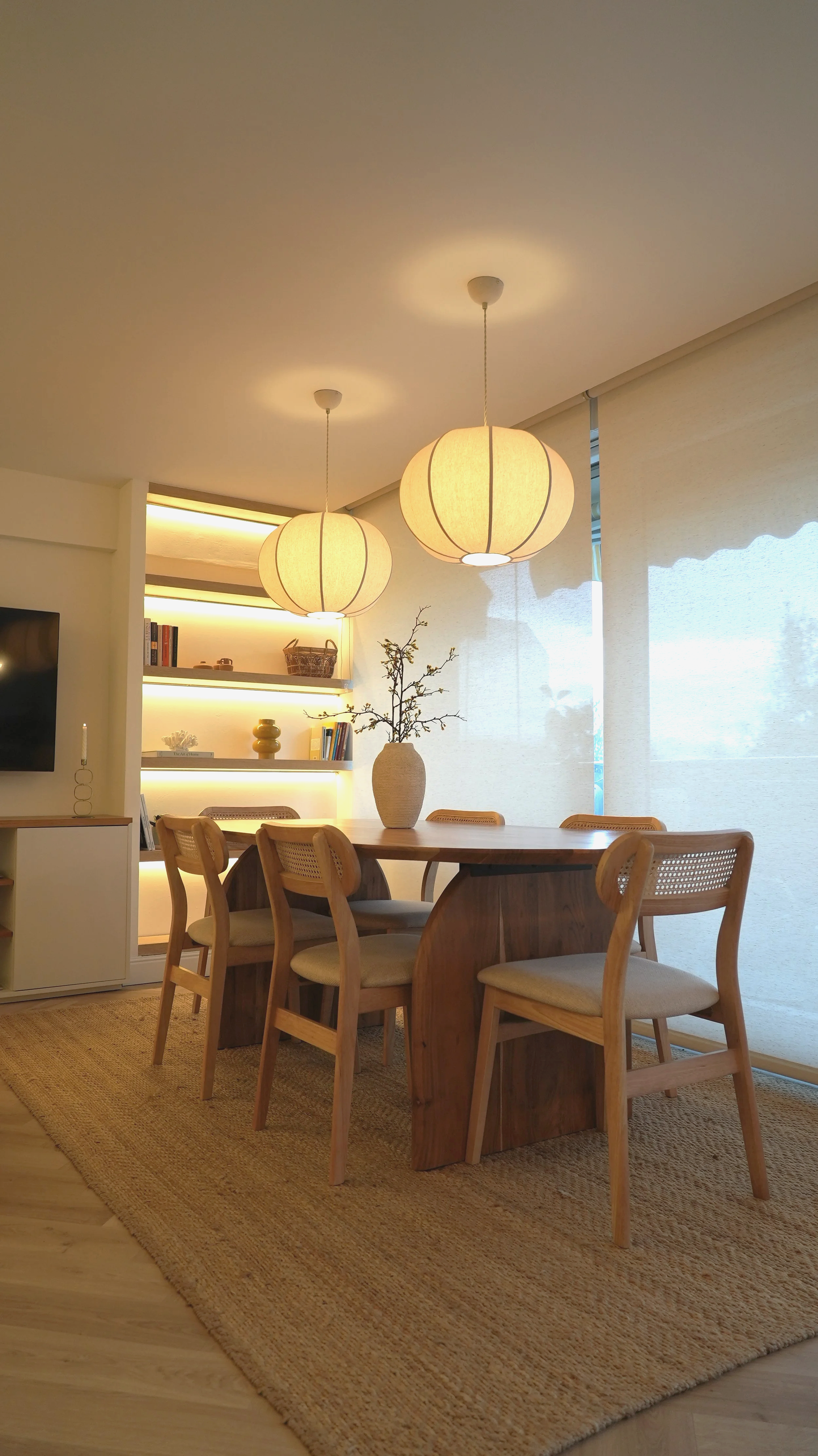 Comedor con mesa de madera y seis sillas, lámparas de papel en el techo, estanterías con libros y objetos decorativos, alfombra de yute, cortinas translúcidas y una vasija con ramas en la mesa.