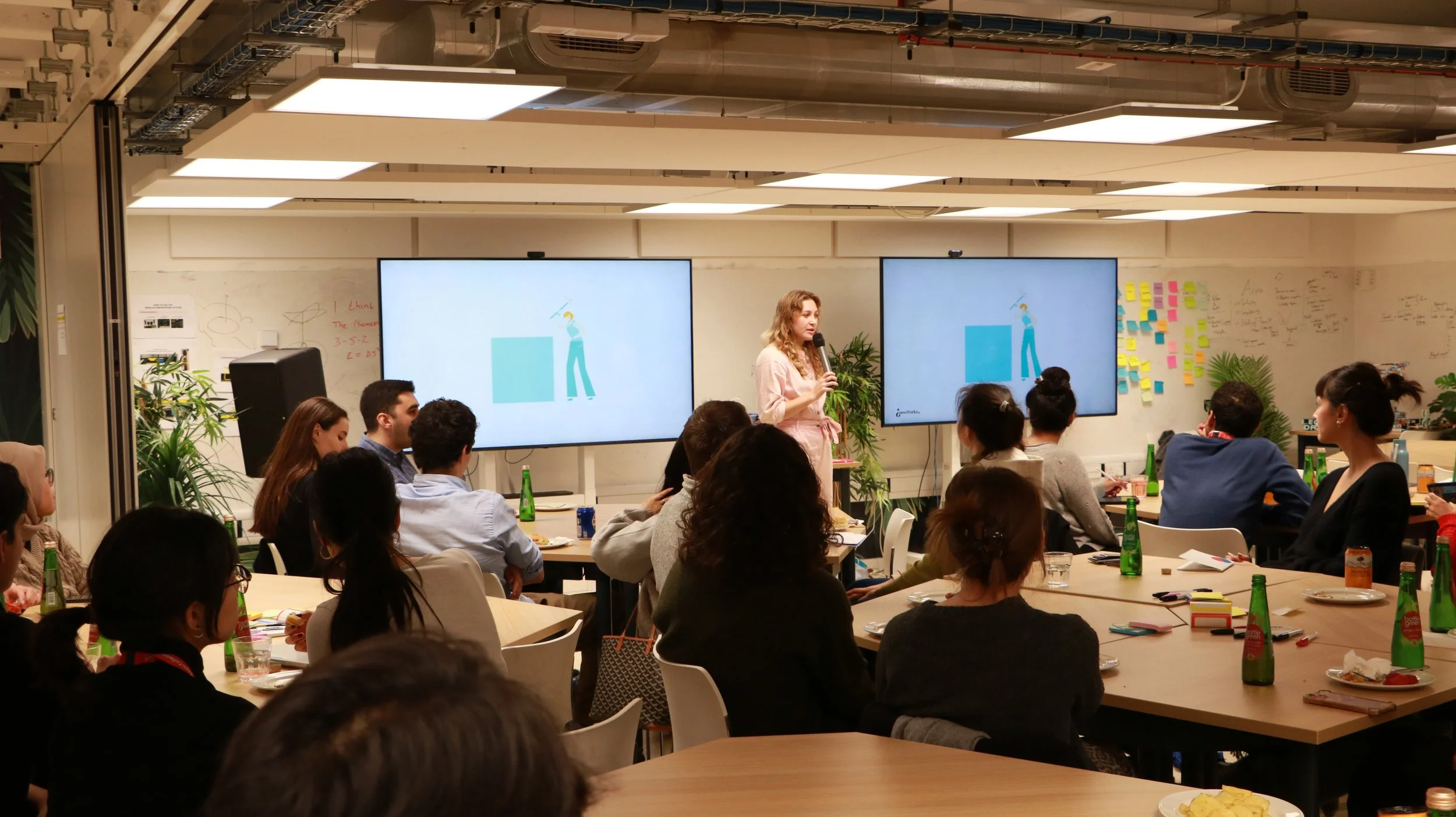 Workshop facilitator presenting to a group of participants in a collaborative training space with whiteboards and screens