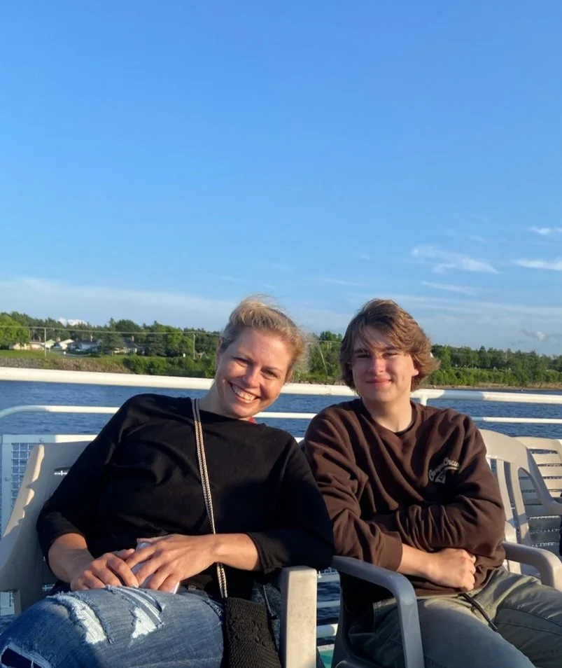 A smiling woman and young man sitting on a bench outdoors near a body of water on a sunny day.