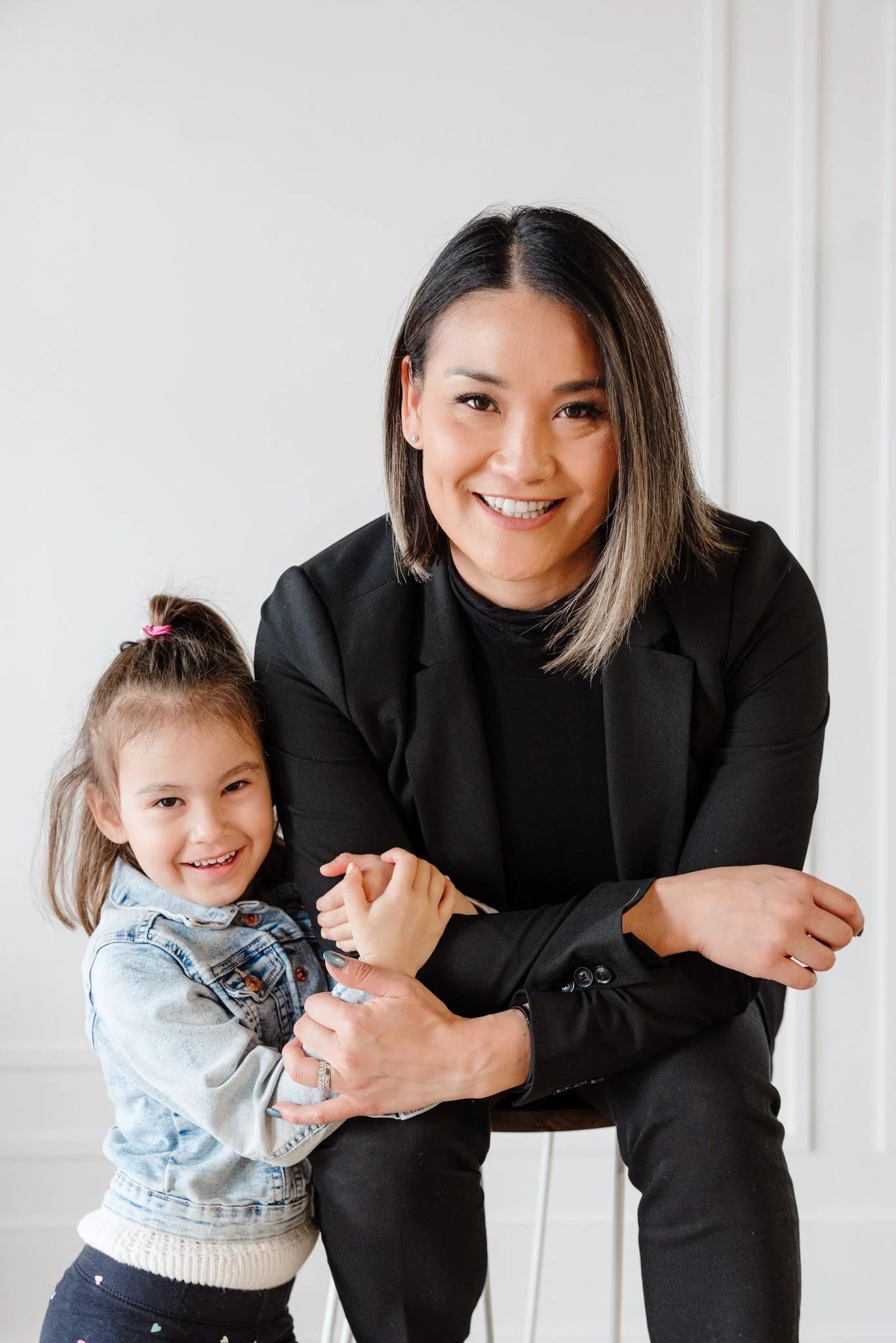 A woman and a young girl smiling, sitting together inside. The woman wears a black jacket and the girl is in a denim jacket and a white sweater, with her hair tied back.