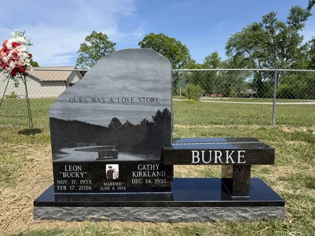 Gravestone with the inscription "Our was a love story" for Leon Bucky Burke and Cathy Kirkland, with their birth and death dates, located in a grassy outdoor setting with a chain-link fence and trees in the background.