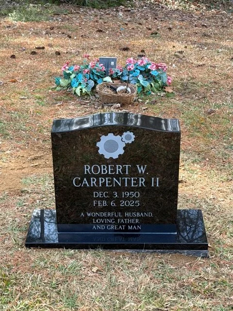 Black granite headstone for Robert W. Carpenter II with a gear and cogs design, displaying his birth and death dates, surrounded by pink and blue flowers and greenery at a gravesite.