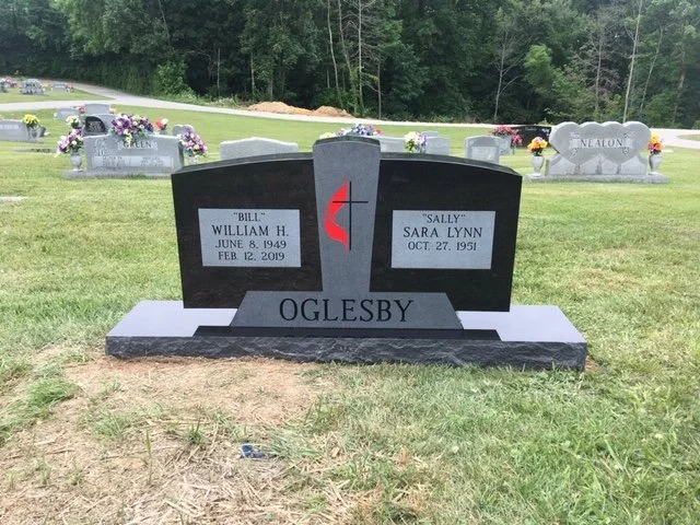 Gravestone in a cemetery with the family name Oglesby, featuring two smaller plaques for Bill William H. and Sally Sara Lynn, with trees and other gravestones in the background.