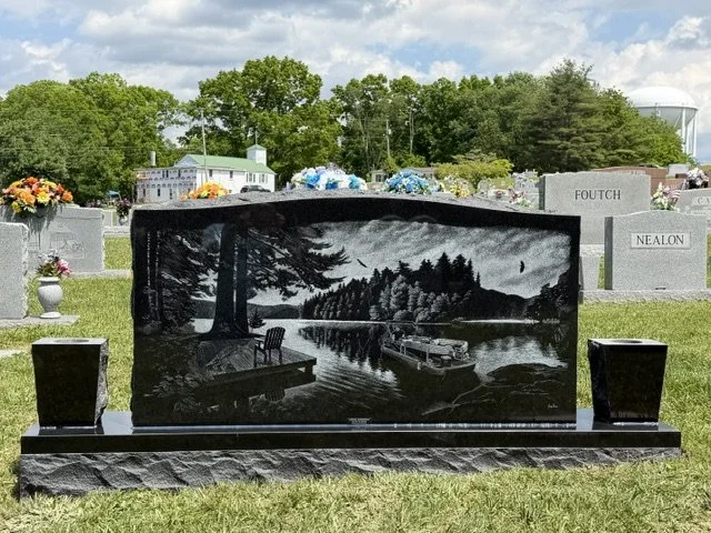 Black polished gravestone with a carved landscape scene of trees, a river, a boat, and a bench, situated in a cemetery. Other headstones and trees are visible in the background.