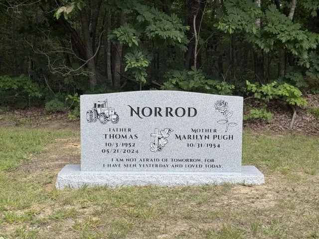 Gravestone for Norrod with images of a tractor, a rose, and a cross, located in a grassy area with trees behind it.