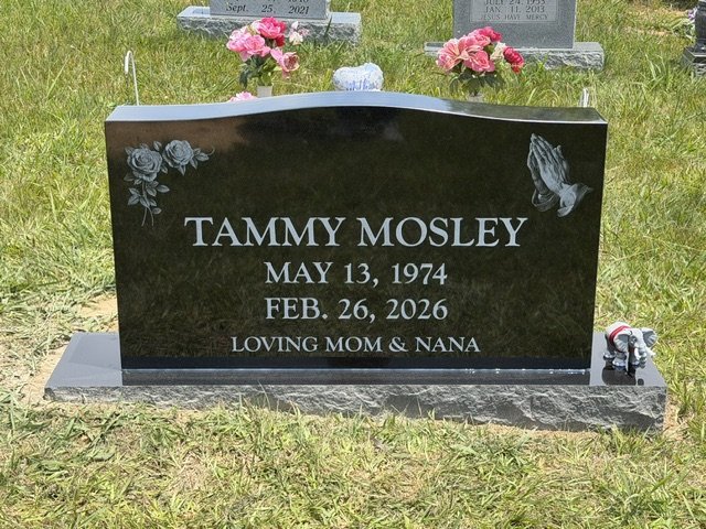 Black granite headstone for Tammy Mosley with embossed roses and a praying hands engraving. It includes her birth and death dates, May 13, 1974, and Feb. 26, 2026, with the inscription 'Loving Mom & Nana.'