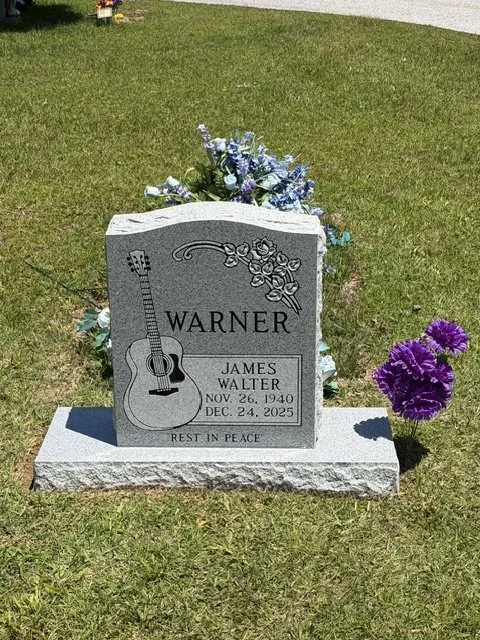 Gravestone with floral arrangements in a grassy cemetery. The tombstone is engraved with the name Warner, a guitar, and the name James Walter, with birth and death dates, and the phrase 'Rest in Peace.'
