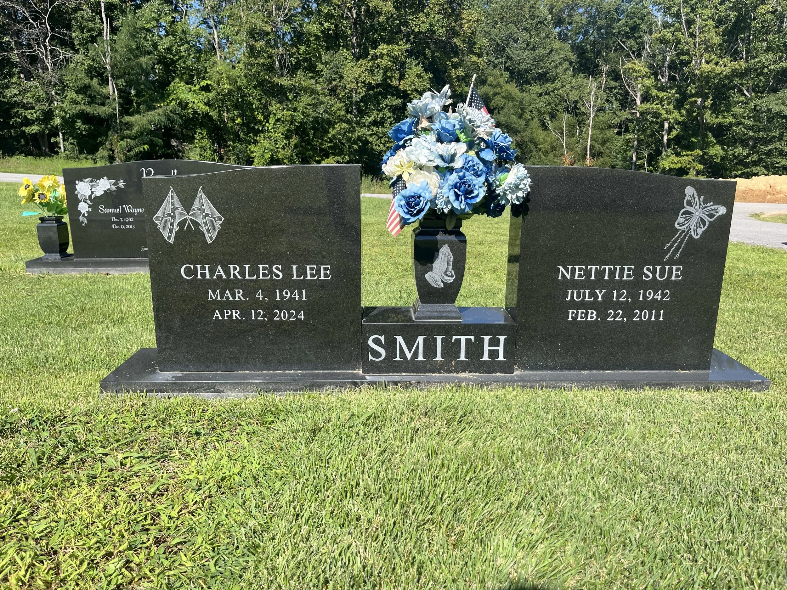 Gravestone with a bouquet of blue, white, and yellow flowers and an American flag, situated in a grassy area with trees in the background.