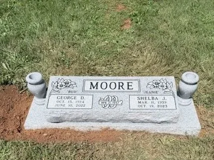 Gray granite gravestone with the name Moore, with inscriptions for George D. and Shelba J., and decorative floral engravings, located in a grassy area.
