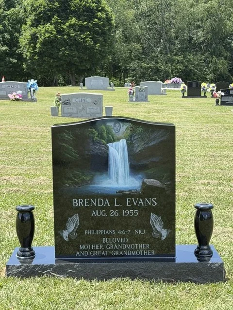 Gravestone in a cemetery with a picture of a waterfall and hands in prayer, inscribed with the name Brenda L. Evans, born August 26, 1955, and a Bible verse. There are other gravestones in the background and trees beyond.