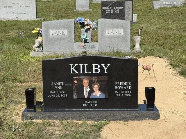 Gravestone of Kilby family with a photograph of a couple, Janet Lynn and Freddie Howard, along with their birth and death dates. The gravestone is black with white lettering, set in a cemetery with other headstones in the background.