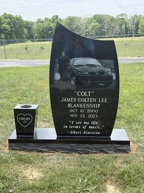 A black memorial headstone for James Colten Lee Blankenship, nicknamed 'Colt,' with a photograph of a man standing next to a car. The headstone has an inscription with his birthdate October 10, 2000, and the date of passing November 22, 2023. The quote 'I see my life in terms of music.' by Albert Einstein is engraved. A small white block with a heart-shaped symbol and the words 'Forever 23' is on the left side of the base.