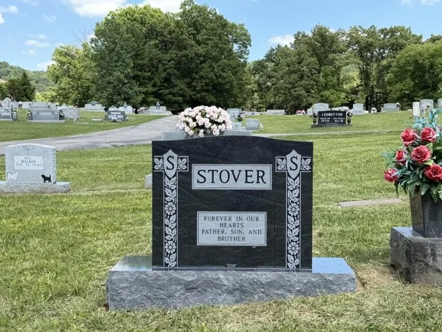 A gravestone in a cemetery with the name 'Stover' and an inscription 'Forever in our hearts, Father, Son, and Brother.' It is surrounded by other graves, flowers, and greenery.