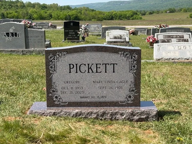 Gravestone with the surname Pickett, for Gregory and Mary Linda Cagle, with birth and death dates, located in a cemetery with other gravestones and green landscape and hills in the background.