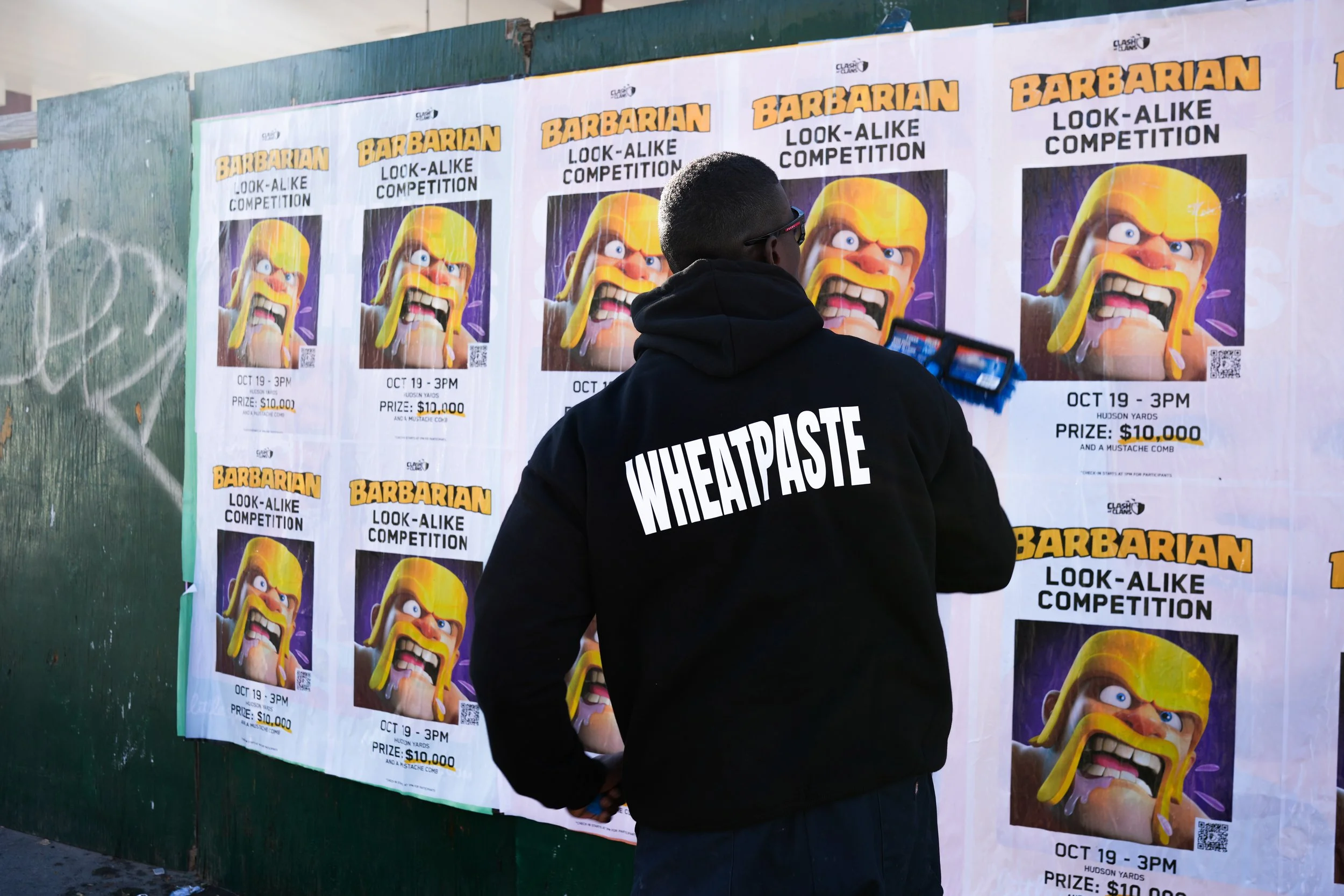 A person with a black hoodie labeled 'WHEATPASTE' on the back, standing in front of a wall of posters for a 'Barbarian Look-Alike Competition.' The posters feature an animated character with long blonde hair and a fierce expression, with event details including the date, time, location, and prize money.