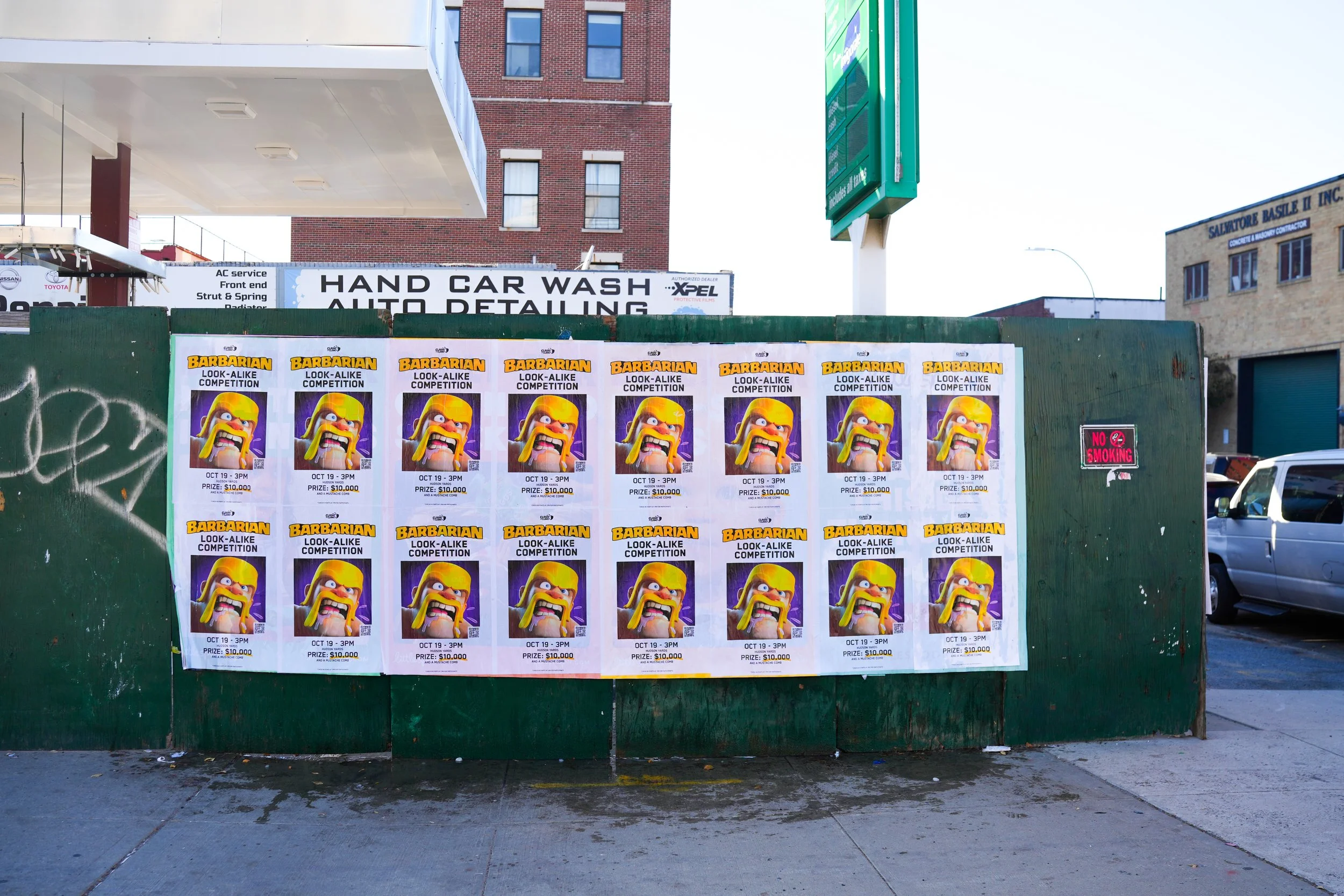 Street scene featuring multiple posters advertising a 'Barbarian Look-Alike Competition' scheduled for October 19 at 3 PM, with a prize of $10,000. The posters display a cartoon-style Viking character with a long yellow beard and helmet, set against a purple background, repeated multiple times on a green construction barrier. In the background, there is a hand car wash and auto detailing shop, a building with a sign, and several parked cars.
