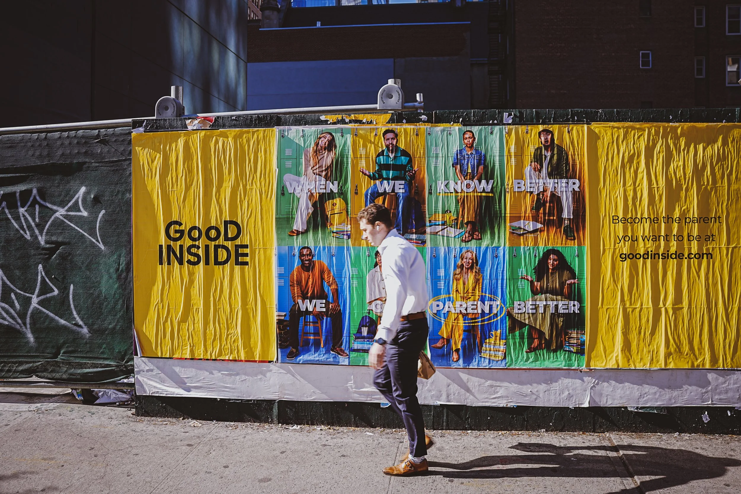 A man walks past a yellow poster on a construction fence with an advertisement about parenting and education. The poster features images of diverse adults and children with text promoting a website goodinside.com for becoming a better parent.