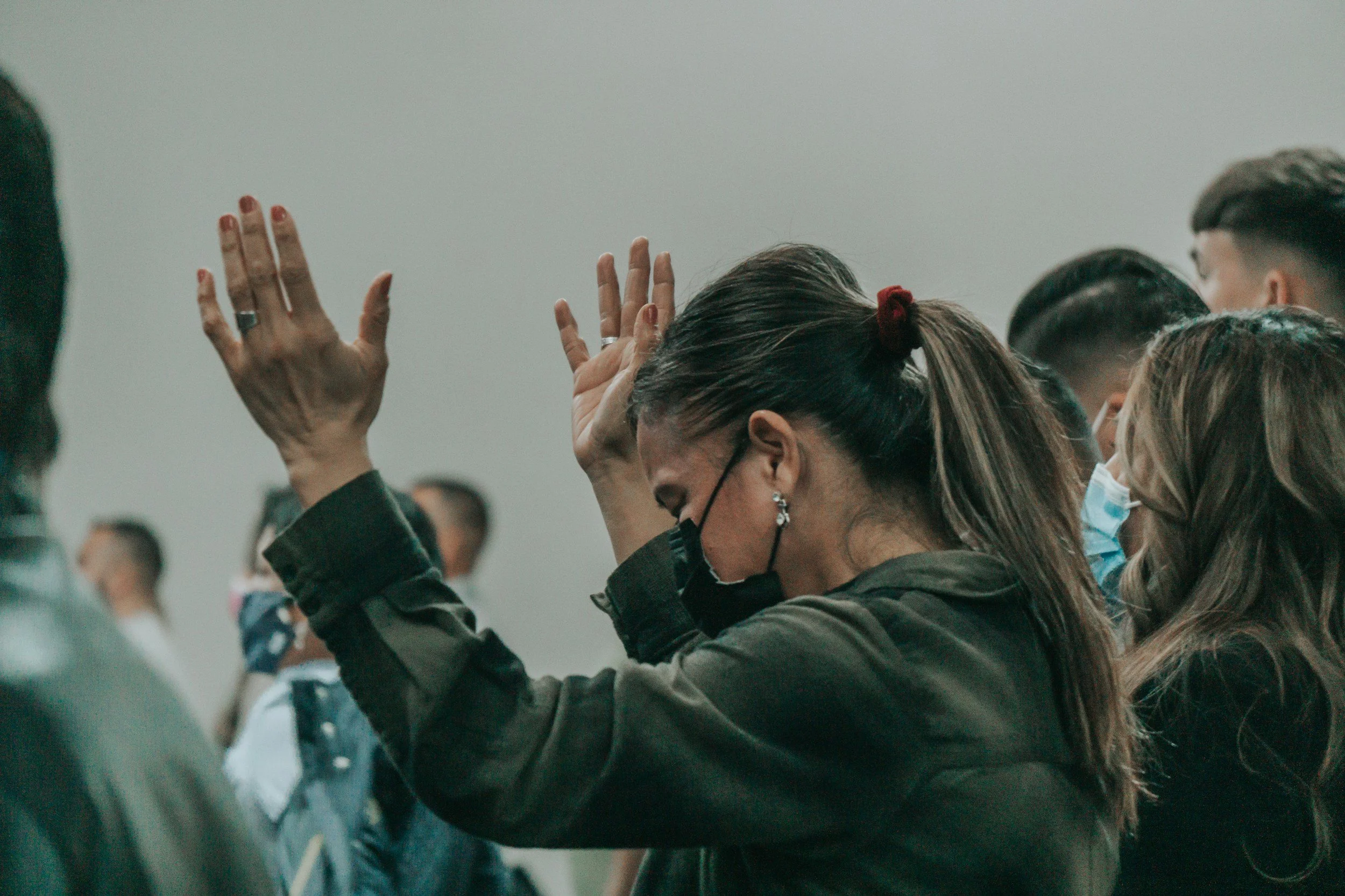 Group of people in an indoor setting, some with eyes closed, one woman with a ponytail and glasses wearing a face mask, raising her hands with her eyes closed.