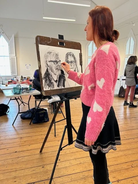A woman in a pink sweater with heart patterns sketches a portrait on a drawing board in an art studio.