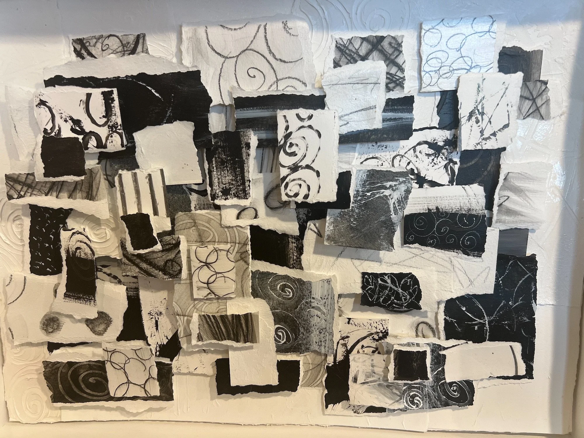 Mixed media collage artwork featuring torn paper pieces, mainly in black, white, and gray with various abstract patterns and scribbles, arranged on a textured white background.