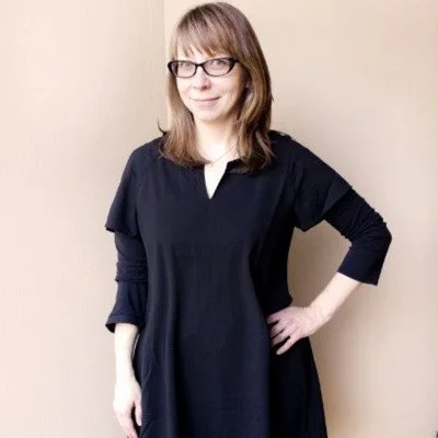 A woman with shoulder-length light brown hair, glasses, and a black dress standing against a beige wall, smiling with one hand on her hip.