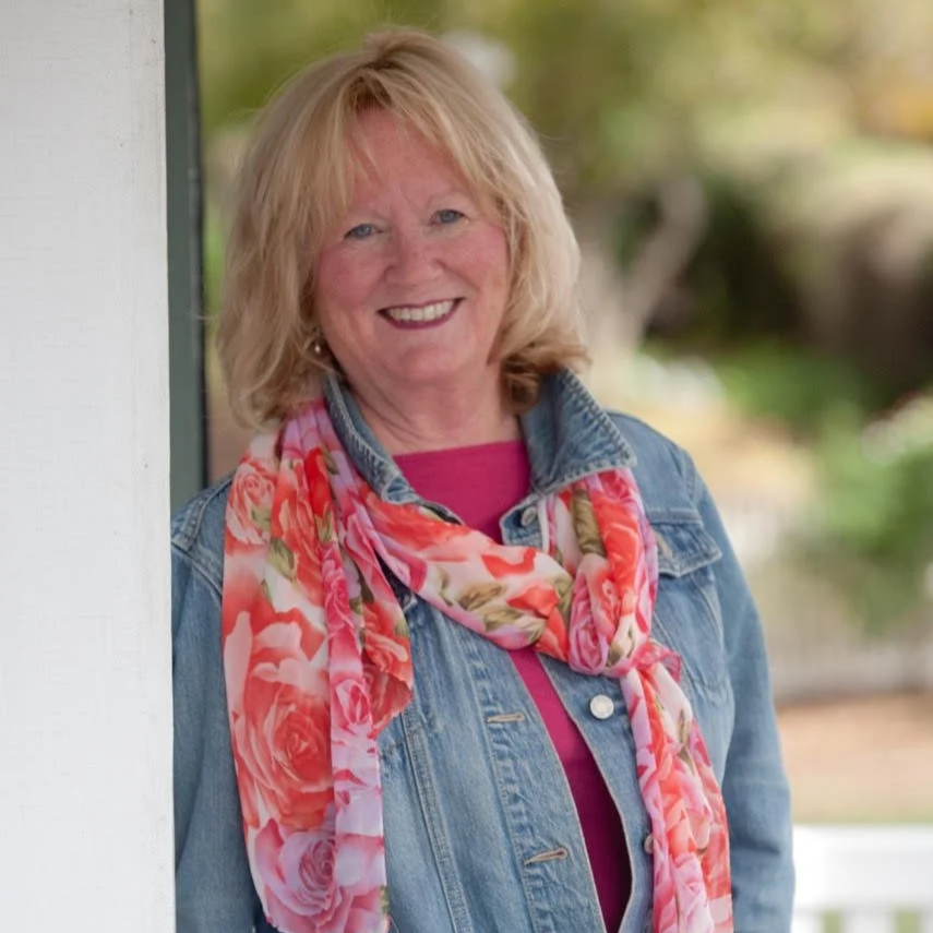 A smiling middle-aged woman with blonde hair standing outdoors, wearing a denim jacket and a colorful floral scarf.