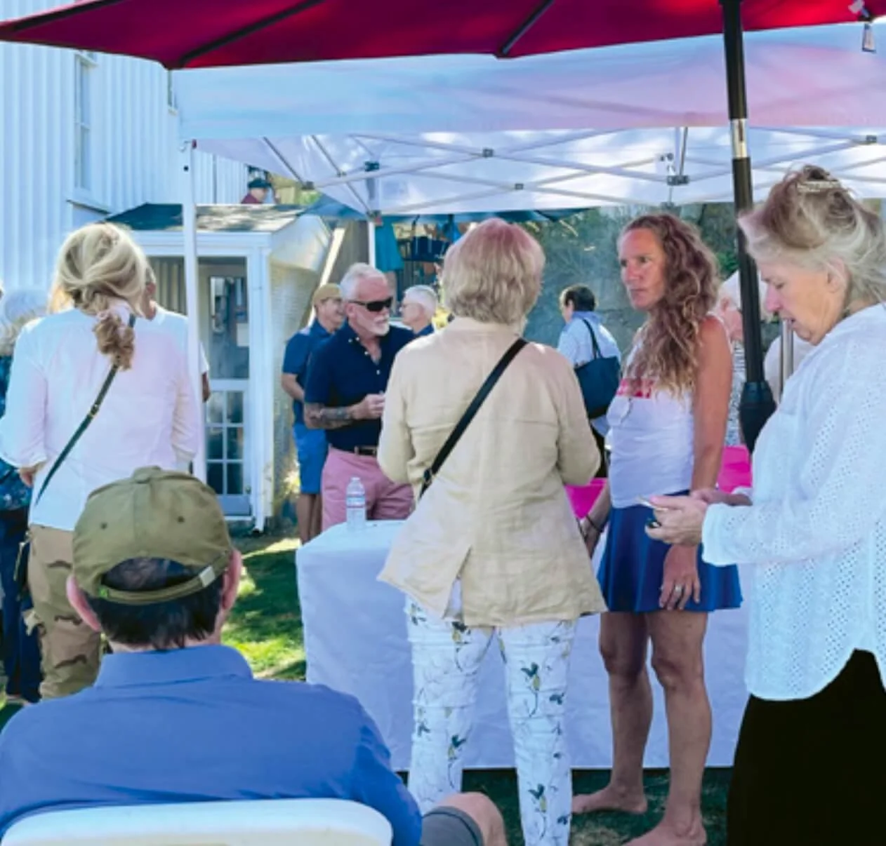 People gathered outdoors under umbrellas in a casual social setting, engaging in conversation. There are women, men, and a child visible, with a white building in the background and tables set up. It appears to be a community or private gathering during daytime.