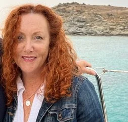Woman with curly red hair smiling, outdoors near water with rocky landscape in the background.