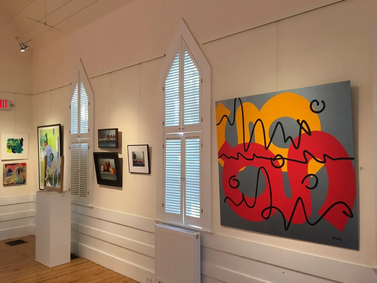 Art gallery wall depicting various colorful paintings and photographs, with a large abstract painting featuring red, yellow, gray, and black colors, and a window with white shutters in the background.