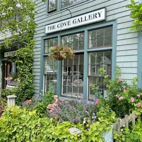 The exterior of The Cove Gallery, a building with light blue wooden siding and large windows, surrounded by lush greenery and flowers.