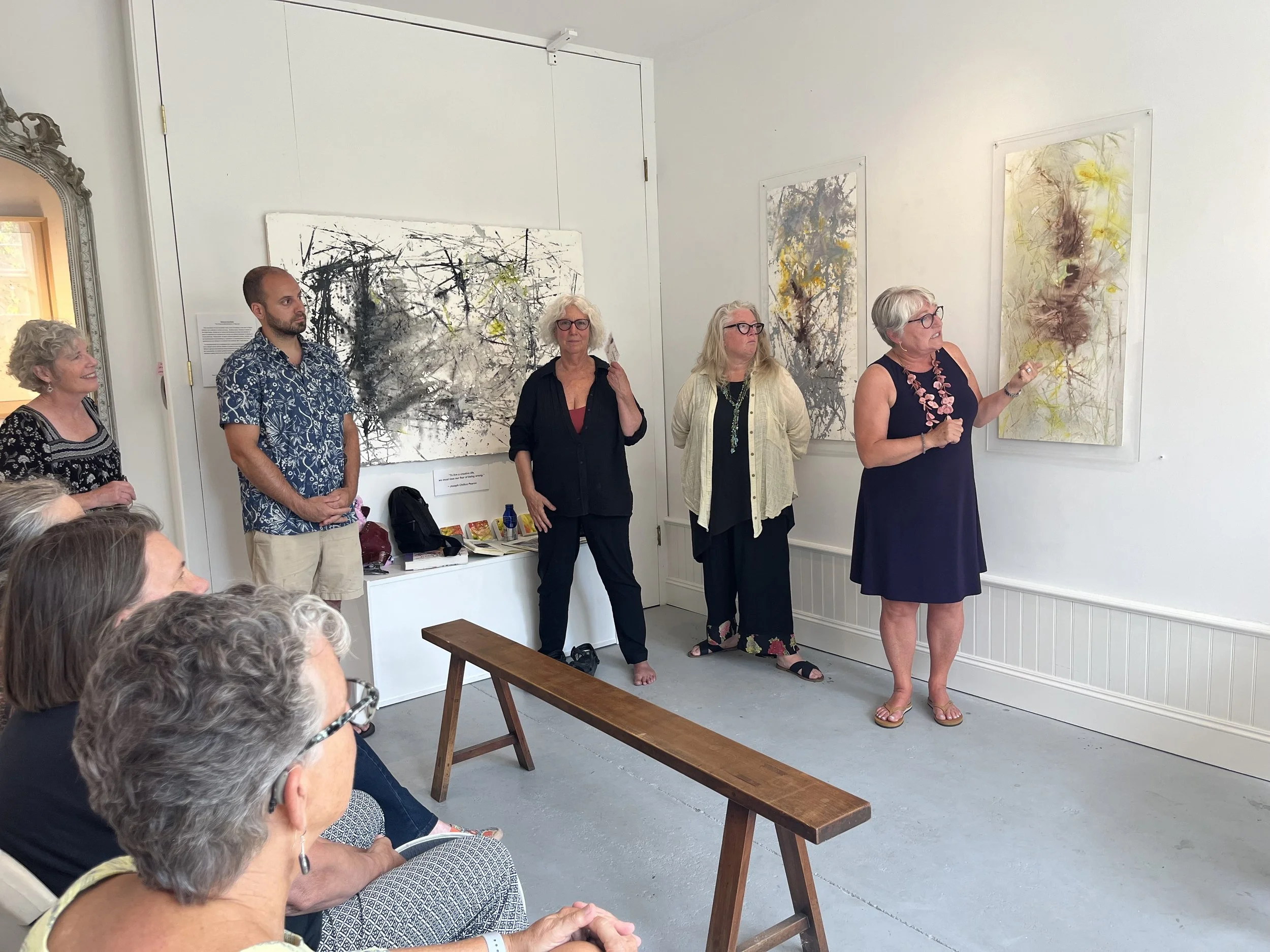 An art gallery opening with five women standing near abstract paintings, one woman speaking or explaining, and an audience seated watching.