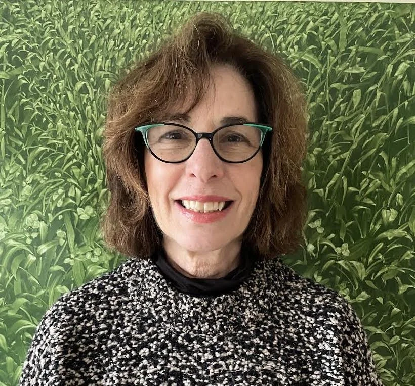 A woman with shoulder-length brown hair, glasses, and wearing a black and white patterned top, standing in front of a green leafy background.