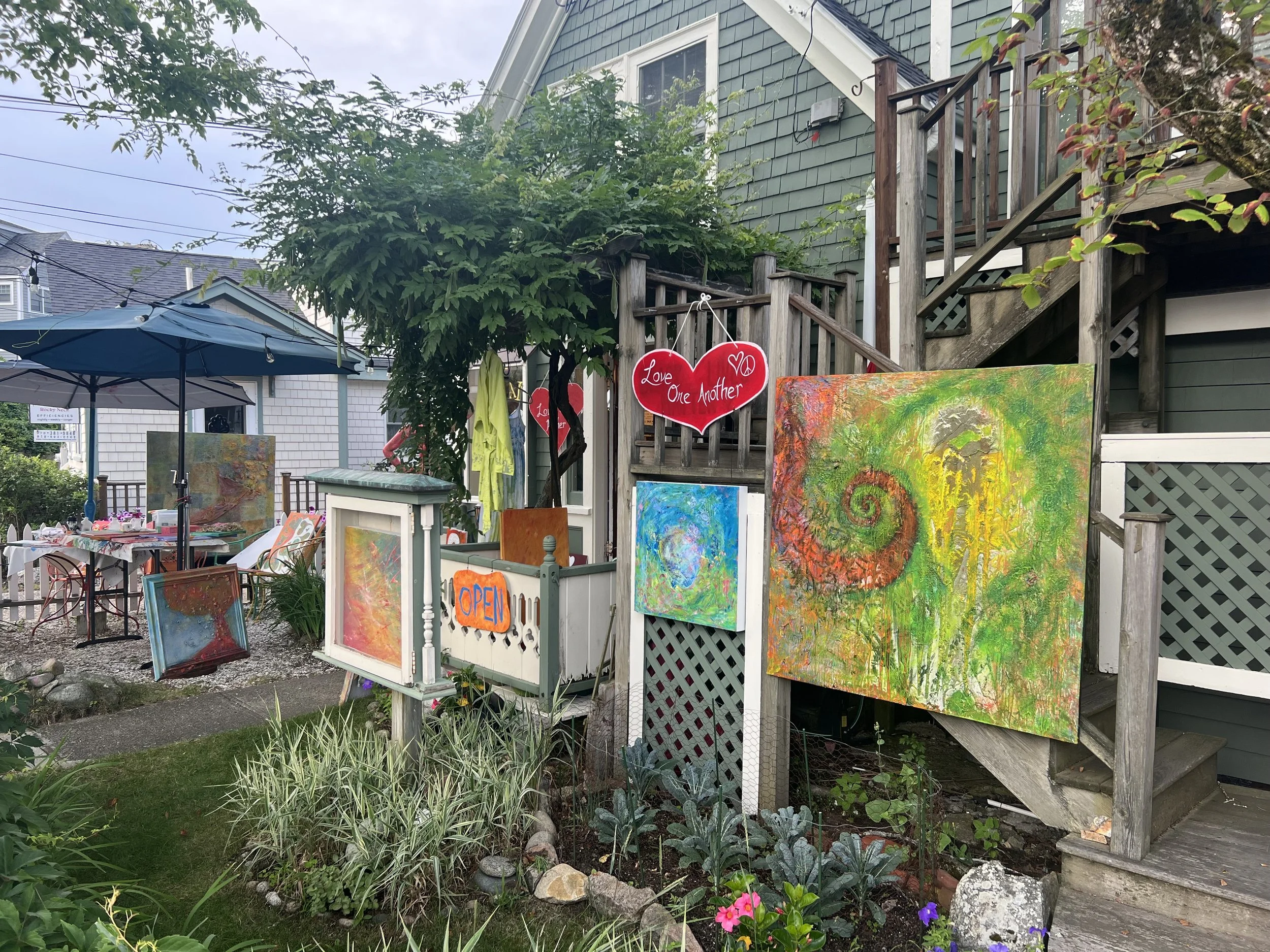 An outdoor art display at a small shop or studio, featuring colorful abstract paintings on various sized canvases, a sign with an orange background that says 'OPEN', and pink heart decorations with the words 'Love One Another'. There is a table with chairs, a green umbrella, and lush greenery including a small tree and flowering plants.