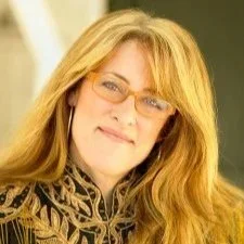 A woman with long blonde hair and glasses, wearing a patterned scarf, smiling softly at the camera.