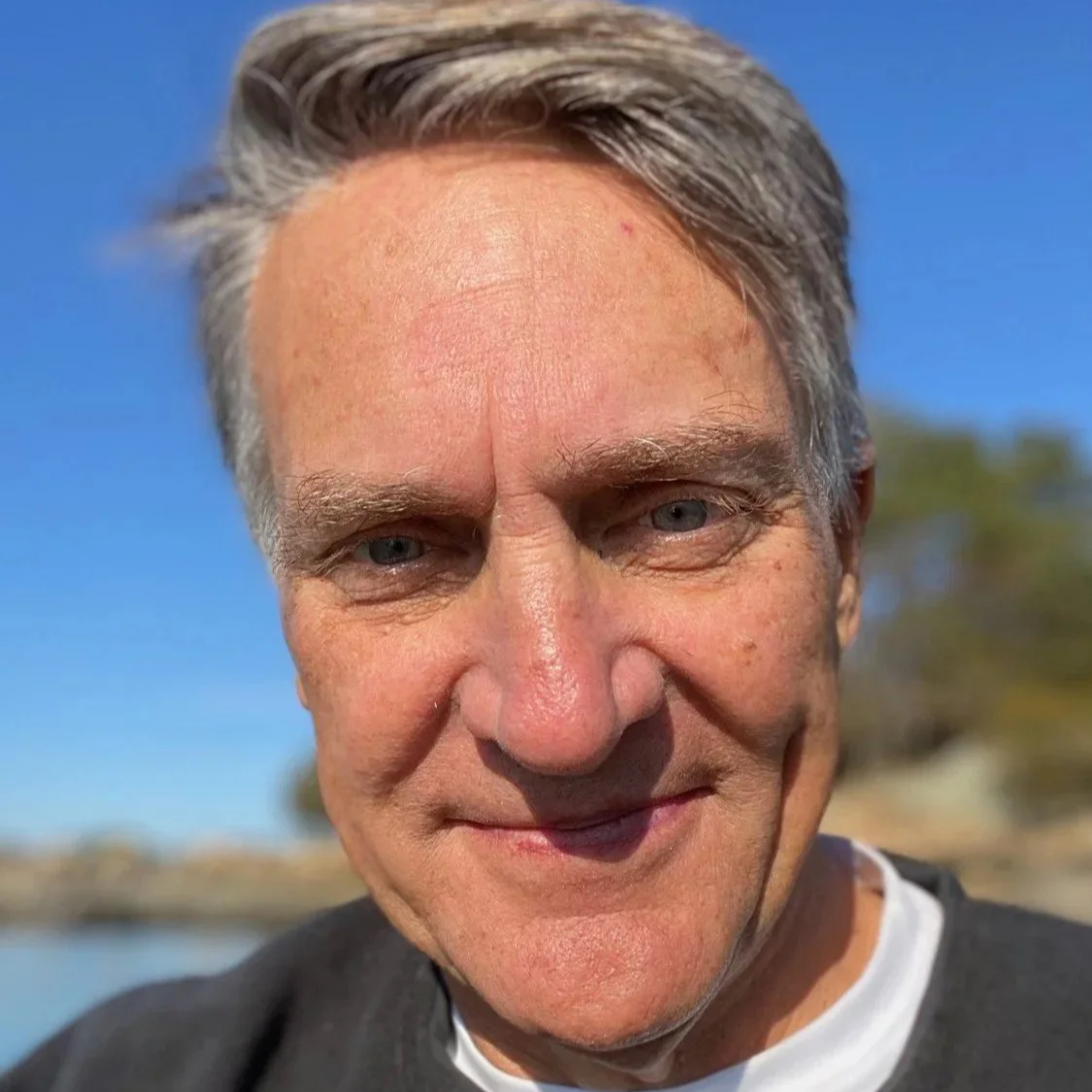 A close-up of a middle-aged man with gray hair outdoors, smiling with a background of blue sky and trees.