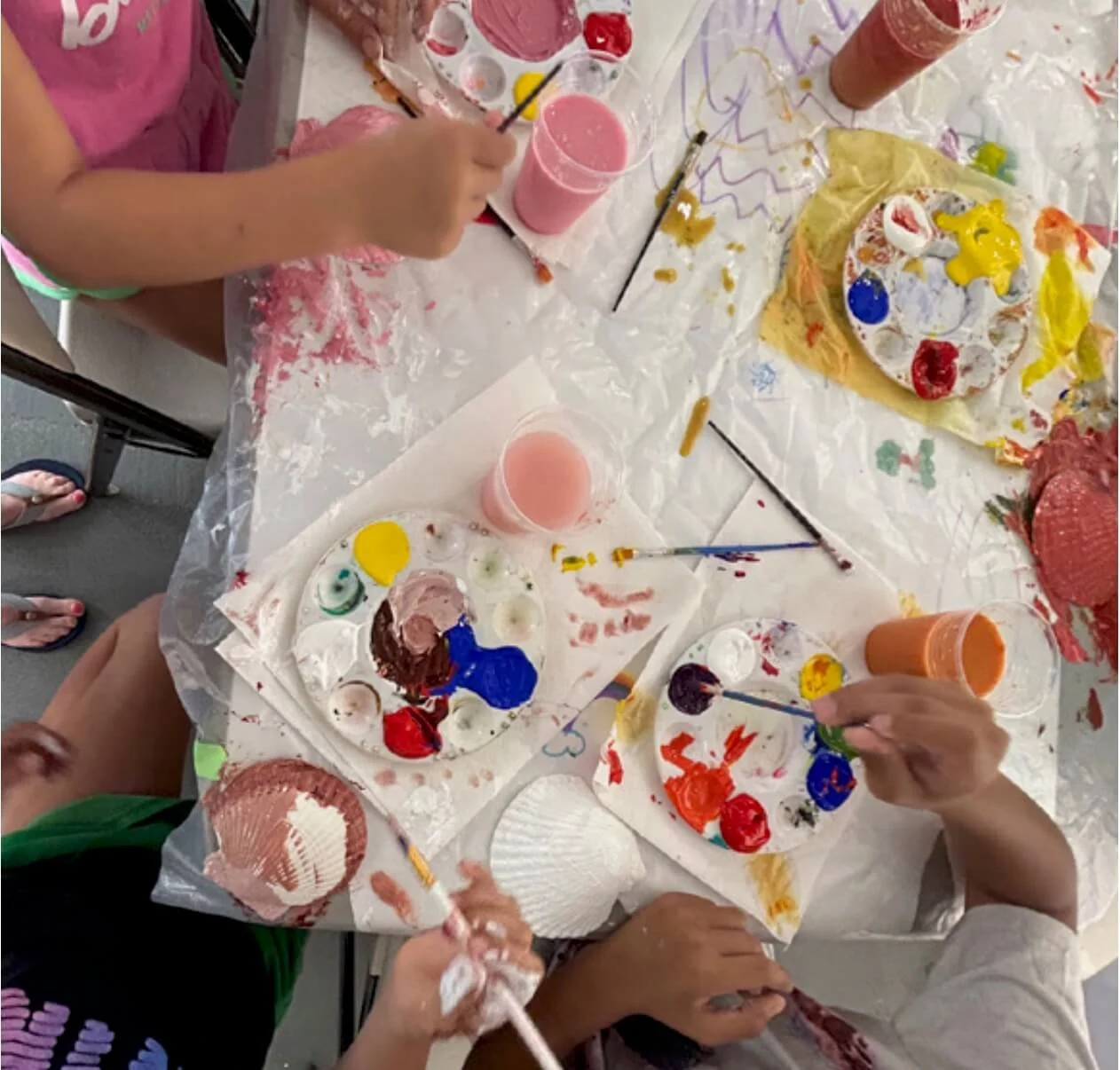 Children engaging in a messy art activity, painting and mixing colors on paper plates and a crepe with paintbrushes and cups of pink paint, surrounded by a covered table with paint splatters.